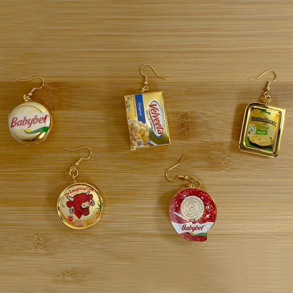 Mini gold Babybel, Velveeta, Babybel Swirl, Laughing Cow, and Leerdammer cheese earrings on a wood backdrop.