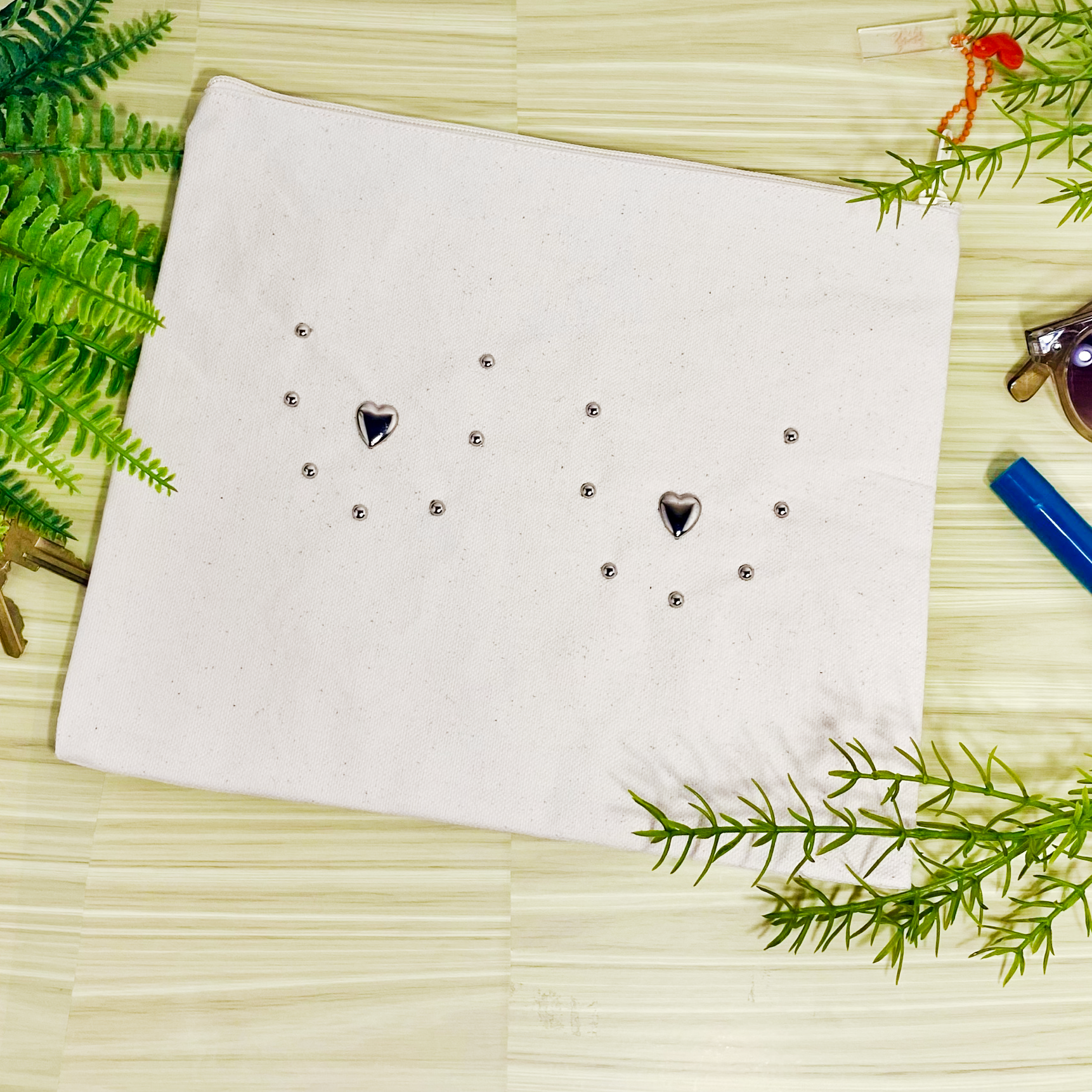  Canvas pouch bedazzled with silver metal studs to outline the shape of breasts with metal hearts for the nipples. The pouch is on a wooden background surrounded by faux plants.